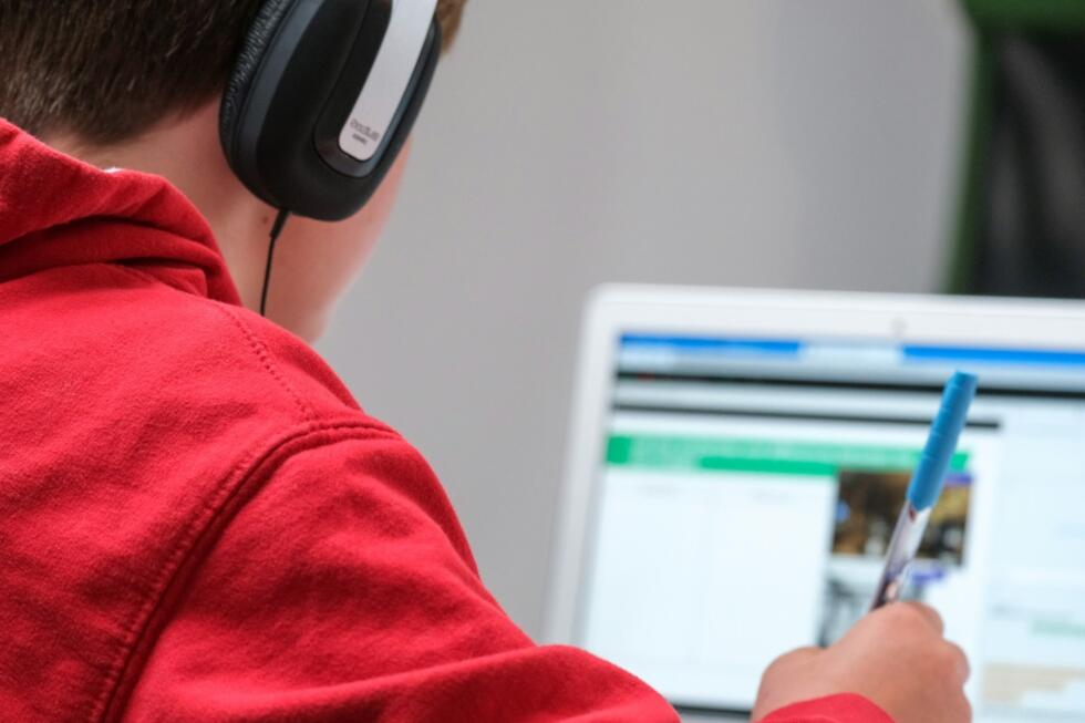 Uczeń ze słuchawkami na uszach notuje podczas nauki z wykorzystaniem laptopa. Unsplash: Compare Fibre