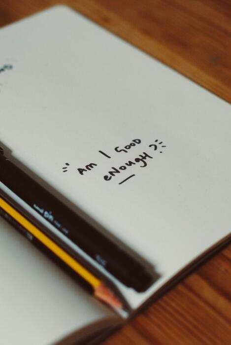 Otwarty notes z napisanym zdaniem "Am I good enough" oraz długopisem i ołówkiem.